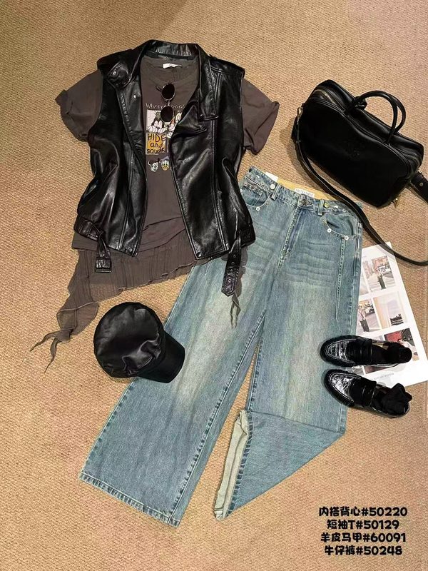 A black leather vest, a grey printed T-shirt, wide light-blue jeans, a black cap, a black bag, sunglasses and black loafers laid out on a beige rug